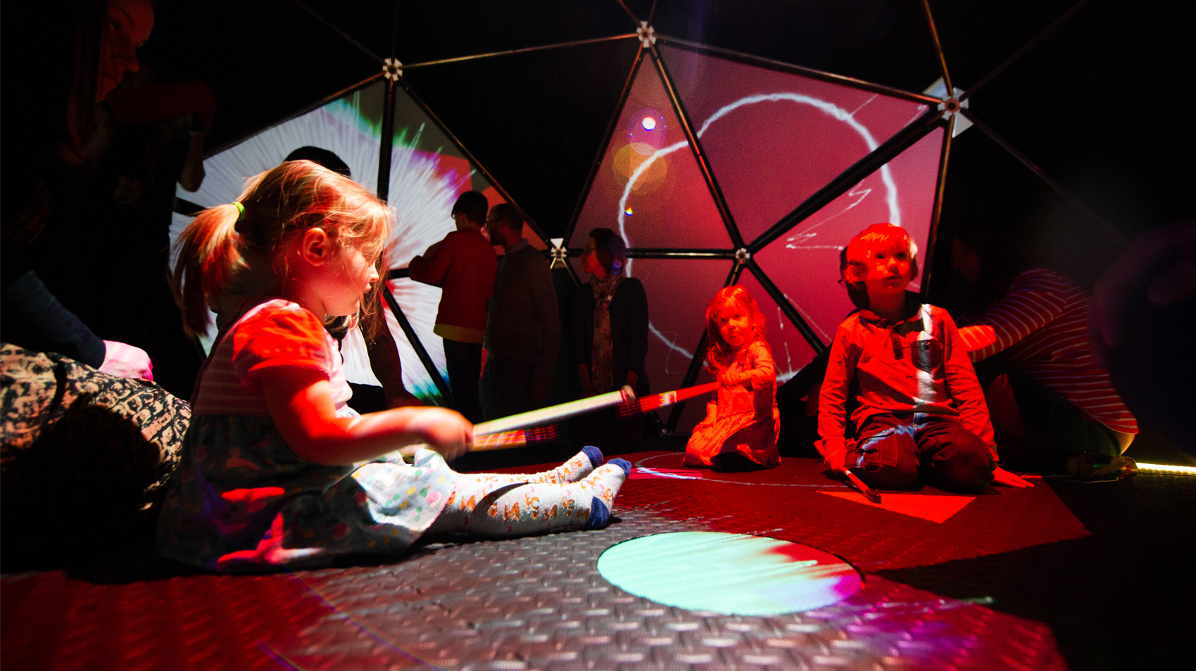 Children play with instruments in dimly lit red light inside a geometric dome
