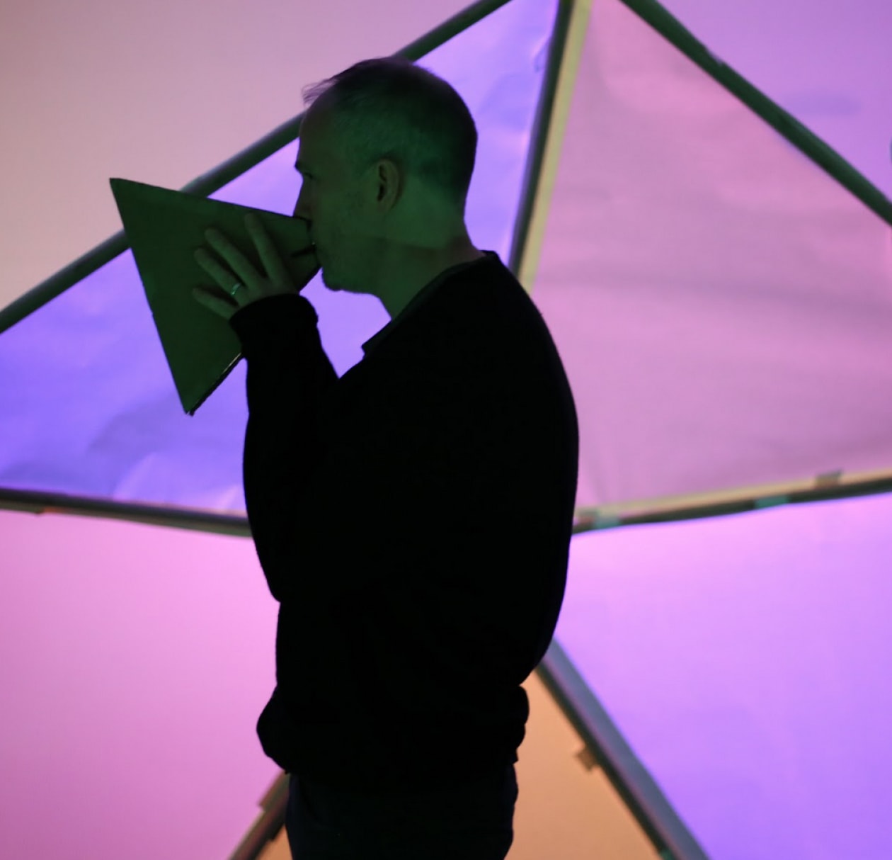 A man speaks through a triangular megaphone infront of purple and pink geometric panels