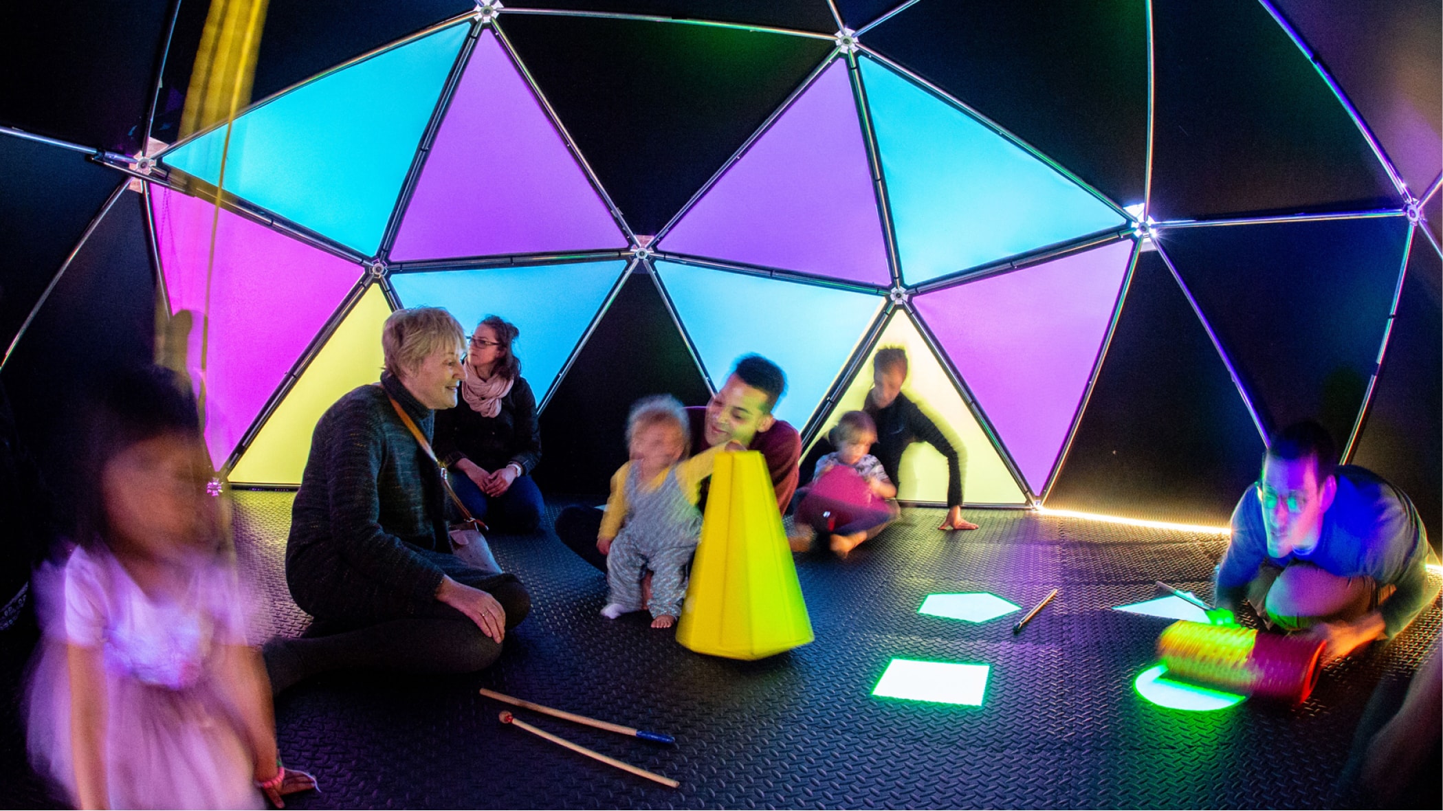 A group of children and adults play with instruments in a brightly lit and colourful geometric dome