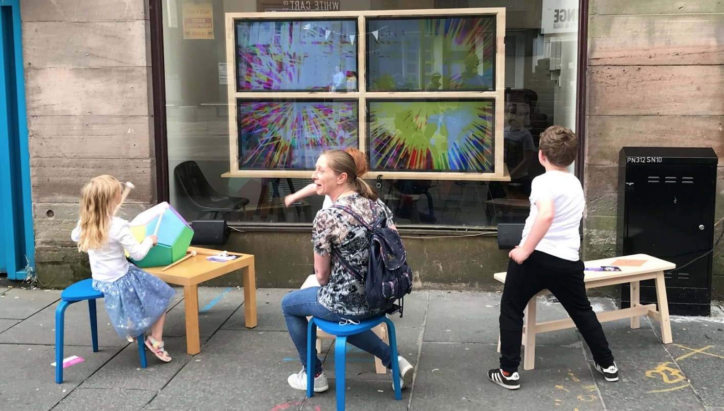 A group of children adults sit on benches and chairs playing with instruments in front of a window with TVs behind the window showing bright colours